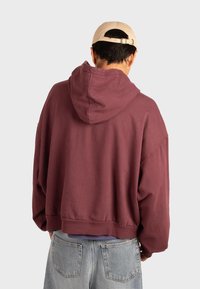 Felpa con cappuccio bordeaux dal taglio morbido, caratterizzata da un cappuccio pronunciato, orlo a coste e spalle basse, abbinata a jeans azzurri chiari.