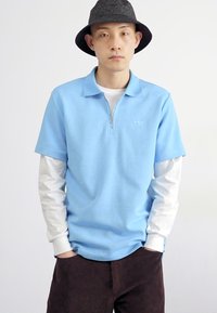 Camisa polo azul clara com colarinho com fecho e tecido texturizado, usada sobre uma camisa de manga longa branca. Calças de napa castanhas. Chapéu preto.