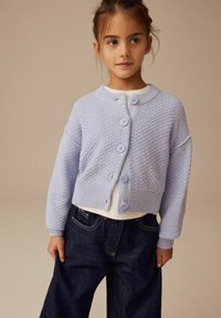 Helllila Strickjacke mit strukturiertem Muster, Rundhalsausschnitt und fünf passenden Knöpfen, kombiniert mit einem weißen Oberteil und dunklen Jeans.