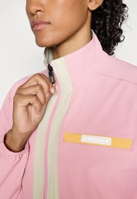 Persona con cabello rizado cerrando una chaqueta rosa y beige con un logo naranja y blanco de "Icepeak" en el pecho.