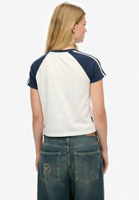 Camiseta de manga corta blanca y azul marino con mangas raglán, que presenta rayas en los hombros en contraste y un dobladillo cropped. Llevada con jeans de mezclilla azul.