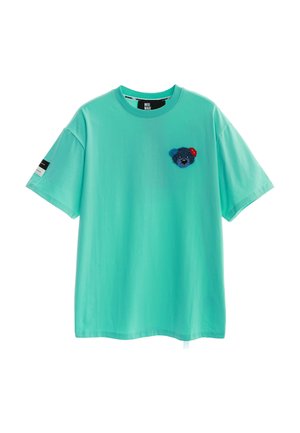 Camiseta de manga corta turquesa con pequeño parche azul de osito de peluche en el lado izquierdo del pecho y una etiqueta en blanco y negro en la manga derecha.