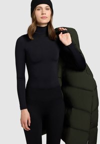 Zwart turtleneck top met lange mouwen gecombineerd met zwarte leggings met hoge taille. Het model draagt een donkergroene gewatteerde jas over haar schouder.