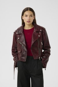 Cropped burgundy faux leather jacket with a crocodile texture, silver hardware, zip closures, and wide lapels. Worn over a red shirt.