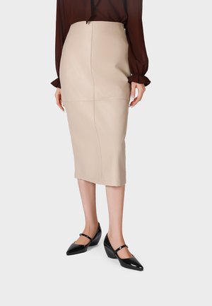 Donna che indossa una gonna a matita beige a vita alta, una maglia scura a maniche lunghe e scarpe Mary Jane nere con punta appuntita, in piedi su uno sfondo semplice.