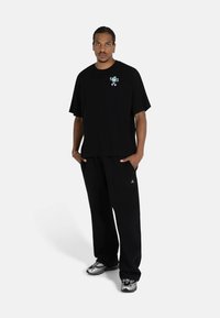 Black cotton t-shirt with a graphic print on the left chest, paired with loose black trousers. Footwear features a silver and black design.