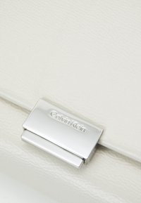 Clutch blanco texturizado con un cierre de metal plateado. Presenta un patrón sutil y el logotipo de la marca en relieve en el cierre.