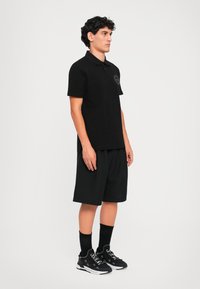Tânăr îmbrăcat cu un tricou polo negru, pantaloni scurți negri, sosete negre și teneși negri cu talpă albă, stând lateral pe un fundal alb simplu.