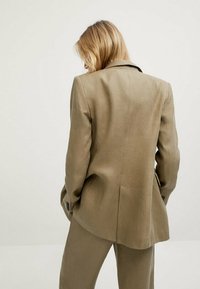 Vaaleanruskea pellavapuvuntakki, jossa on istuva malli, viistot läpät ja tuuletus takaosassa. Teksturoitu kangas ja jämäkkä siluetti.