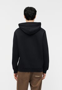 Άτομο που στέκεται με την πλάτη στραμμένη, φοράει μαύρο φούτερ με κουκούλα και καφέ παντελόνι με το λογότυπο PEGADOR στην πίσω τσέπη, μπροστά σε απλό φόντο.