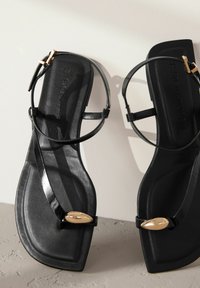 Černé ploché sandály s tenkými popruhy a zlatou ozdobou ve tvaru listu. Hladký povrch a minimalistický design.