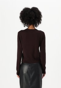 Frau mit lockigem Haar, die einen dunkelbraunen Langarmpullover und einen schwarzen Lederrock trägt, steht mit dem Rücken zur Kamera.