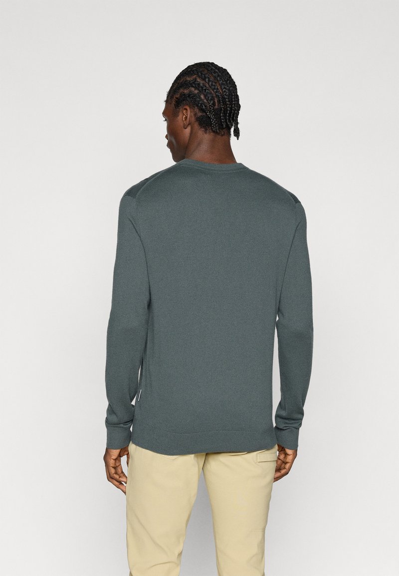 Pull en tricot vert foncé à manches longues, col rond, et bord-côtes aux poignets et à l'ourlet. Fentes sur les côtés et étiquette sur le côté.