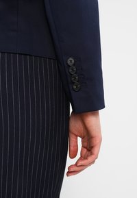 Granatowa marynarka o dopasowanym kroju, z pod spodem pasiastym garniturem oraz pięcioma czarnymi guzikami na rękawie pod gładkim materiałem.