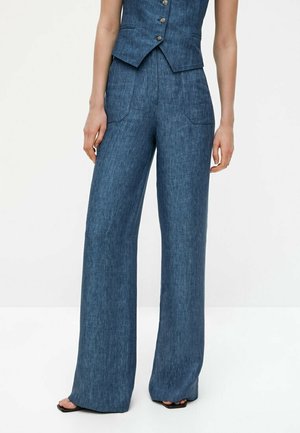 Woman wearing high-waist, wide-leg blue denim pants and matching sleeveless buttoned vest, standing barefoot on white floor.