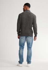 Bărbat întors, îmbrăcat cu un pulover texturat gri, blugi bleu deschis și adidași albi, pe un fundal simplu de culoare deschisă.