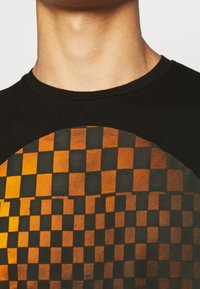 Zwart T-shirt met een ronde graphic met afwisselende oranje en zwarte geblokte patronen. Zachte stof met een klassieke ronde hals.