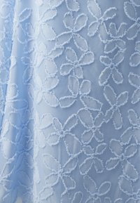 Vaaleansininen kangas, jossa on teksturoitua kukkakirjontaa, jossa on monimutkaisia, pörröisiä terälehtiä ja yksityiskohtaisia ääriviivoja, luoden pehmeän, kerroksittaisen ilmeen.