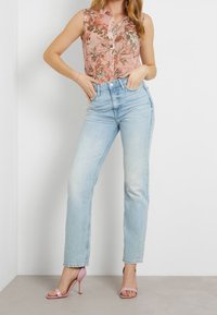 Camiseta sin mangas con estampado floral en rosa con cierre de botones, combinada con vaqueros de talle alto en azul claro. La modelo lleva sandalias de tacón rosas.