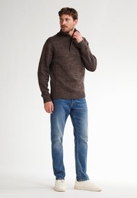 Man die de kraag van een bruin gebreid sweater aanpast, gekleed in blauwe jeans en witte sneakers, staande tegen een effen lichte achtergrond.