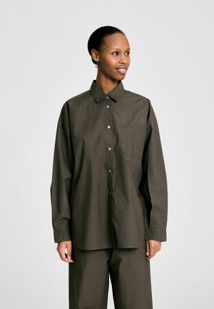 Vrouw draagt een donkerolijfgroene oversized blouse met knopen en een borstzak, gecombineerd met wijde broekspijpen, staand tegen een effen lichte achtergrond.