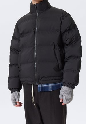 Personne portant une doudoune noire fermée, des gants en tricot gris sans doigts, un pantalon noir, et une chemise à carreaux bleue non rentrée en dessous.