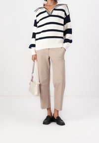 Nainen, jolla on beiget lyhyet housut, mustat loaferit, valko-ruskopallokuvioinen neule ja pieni beige käsilaukku.