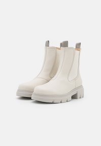 Pár krémově bílých kožených Chelsea bot s robustními gumovými podrážkami a šedými textilními poutky na čistě bílém pozadí.