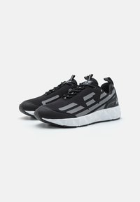 Sorte atletiske sko med en tekstureret overdel, som har grå akcenter og en polstret hvidsål med et geometrisk design. Slanke snørebånd og stropper.