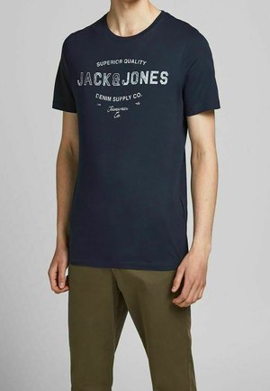 Mand iført navy blå Jack & Jones T-shirt og olivengrønne bukser, stående mod en ensfarvet lys grå baggrund.