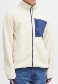 Fleecejacke in Creme mit einer marineblauen, mit Reißverschluss versehenen Brusttasche. Verfügt über einen hohen Kragen und einen Frontreißverschluss, seitliche Taschen und gerippte Bündchen.