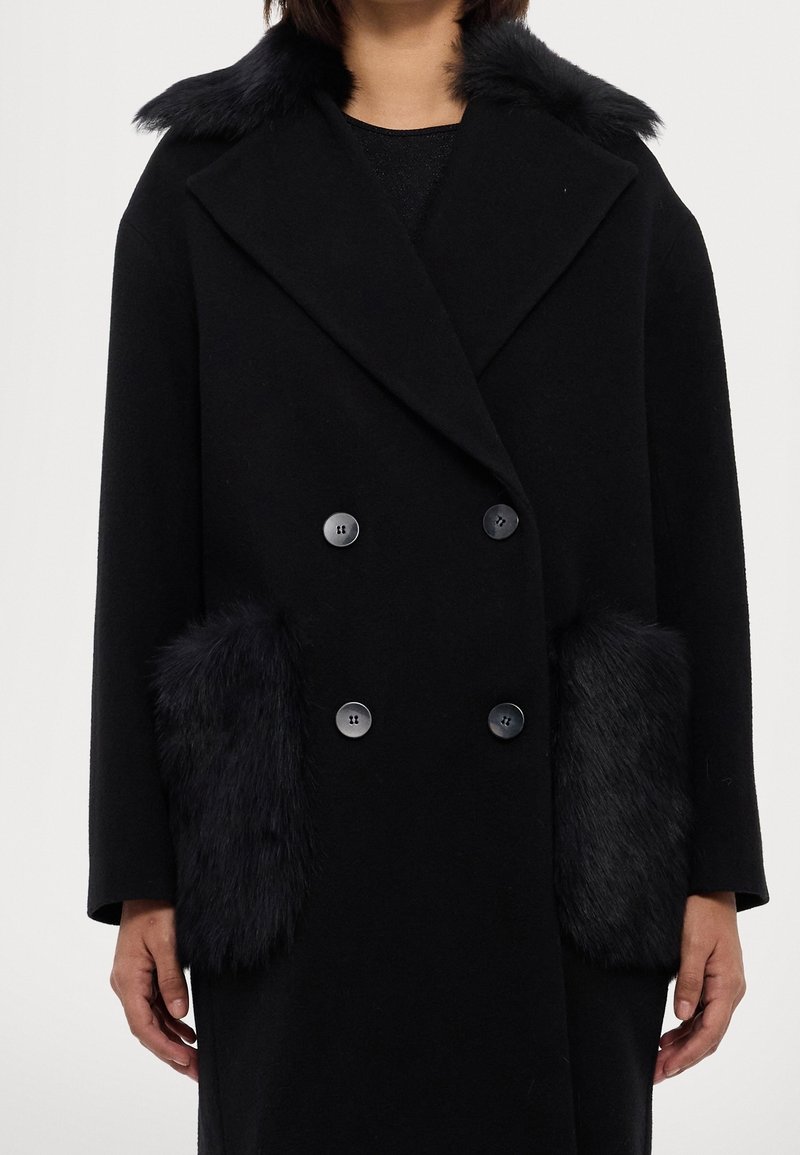 Manteau noir à double boutonnage avec de gros boutons, col en fausse fourrure, et poches plaquées assorties en fausse fourrure, porté par une personne debout, les bras le long du corps.