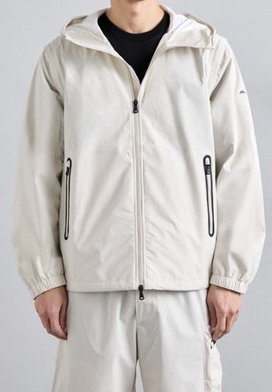 Homme portant une veste à capuche beige clair avec fermeture éclair frontale et poches latérales zippées, associée à un pantalon beige clair assorti.