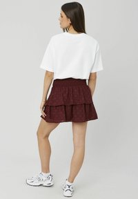 T-shirt blanc à manches courtes associé à une jupe superposée bordeaux avec un tissu texturé et une taille smockée, portée avec des baskets blanches.