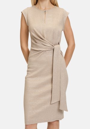 Vrouw draagt een mouwloze beige jurk tot op de knie met glans, een keyhole halslijn aan de voorkant en een geplooide taille strikdetail.