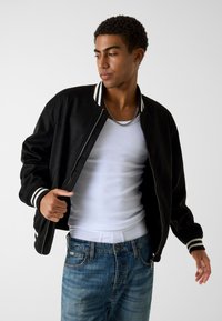 Bomber nero con collo e polsini a costine bianchi a righe, abbinato a una canotta bianca e jeans blu strappati, con zip frontale.