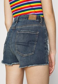 Gros plan d'une personne portant un short en jean bleu foncé avec des bords effilochés et un haut sans manches à rayures arc-en-ciel.