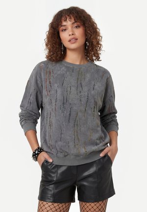 Kvinde med krøllet hår iført en grå slidt sweatshirt, sorte lædershorts, netstrømpebukser og et armbånd med spikes, stående med hænderne i lommerne.