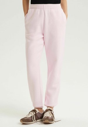 Osoba nosząca jasnoróżowe polarowe spodnie dresowe z elastycznym pasem i kieszeniami, w połączeniu z brązowymi i beżowymi sneakersami na sznurowadła.