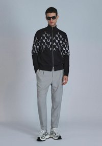 Zwarte zip-up jack met groot wit tekstpatroon, grijze op maat gemaakte broek en zilveren sneakers met zwarte accenten. Model draagt een zonnebril.