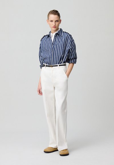 Camisa de botón a rayas en color marino y blanco con adornos en los hombros, combinada con pantalones anchos blancos y zapatos marrones tipo mocasín.