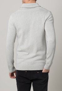 Světlounce šedý pletený svetr se složeným límcem a žebrovaným lemem, nošený s tmavými džínami. Textura působí měkce a pohodlně.