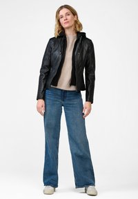 Svart skinnjacka med en fuskpälsfoder, kombinerad med en ljusbeige tröja och vida blå jeans. Modellen står framför en vit bakgrund.