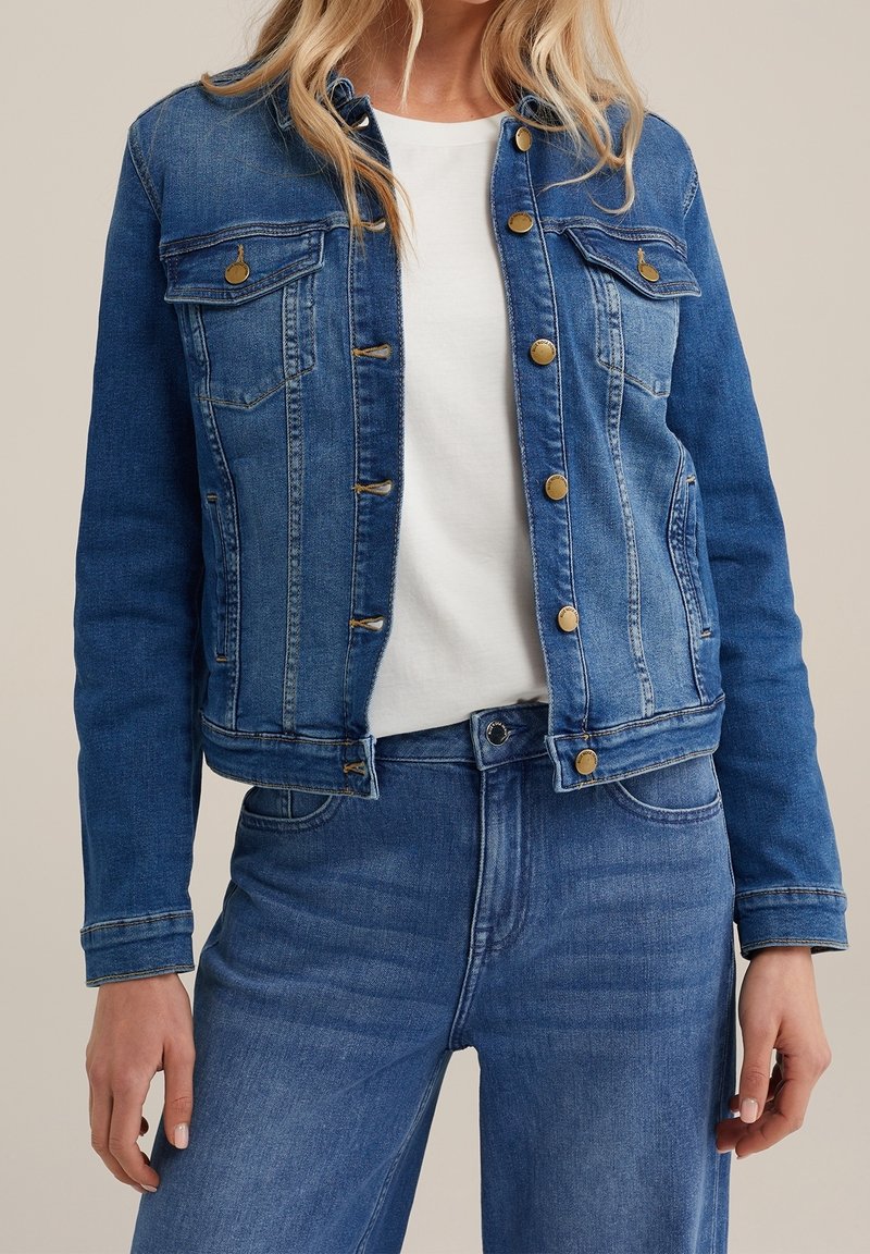 Blå denimjacka med klassisk passform, som har två bröstfickor, guldfärgade knappar och stickningsdetaljer. Bärs över en vit skjorta.