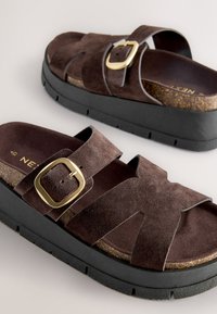 Paire de sandales en daim marron avec larges brides, boucles dorées, semelles en liège et semelles épaisses noires texturées sur une surface claire.