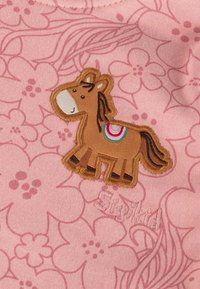 Rosa Stoff mit floralem Muster zeigt ein braun gesticktes Pferd mit einem mehrfarbigen Sattel, neben dem Wort "Siyka", das in Pink gestickt ist.