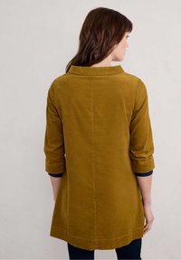 Senfgelbes Corduroy-Tunika mit Dreiviertelärmeln, einem Kragen und geradem Schnitt, die sichtbare Nähte am Saum aufweist.