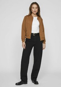 Hnedý bomberský kabát so zipsovými vreckami, biele tričko, čierne nohavice s vysokým pásom a čierne mokasíny. Hladká látka a voľný strih.