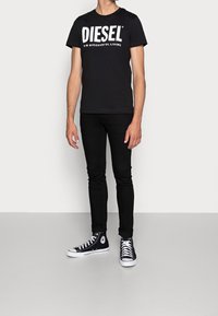 Muž v černém tričku Diesel, černých skinny džínách a černých vysokých teniskách Converse stojí proti jednoduché stěně a podlaze.