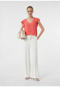 Korallfarbene V-Ausschnitt-Bluse mit gerafften Ärmeln, kombiniert mit hochgeschnittenen weißen weiten Hosen. Das Model hält eine kleine cremefarbene Handtasche.
